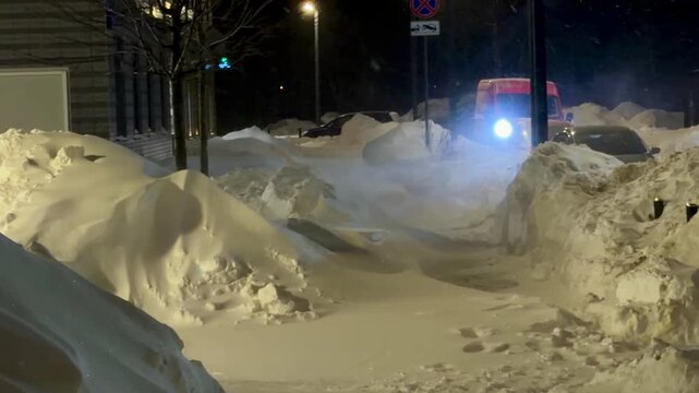 Night city timelapse of extreme snowfall in a residential area. Cars almost completely buried in deep snow drifts on a street with bright headlights of a moving vehicle. Winter blizzard in a city.