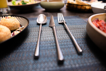Stainless steel cutlery set on blue textured tablecloth with plates of food in background, dining...