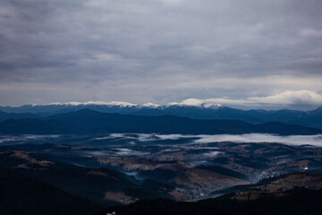 Obraz premium An open panorama shows blue ridges under a dense sky while patches of fog slide across a sprawling valley. Far ranges carry snow and the land below holds scattered villages