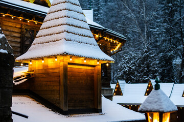 Snow layered turret glows warmly above the rooftops of a wooden complex. Strings of lights sparkle across the scene while a dark forest frames this peaceful mountain village evening. © Oleh