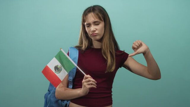 Young mexican student woman holds mexican flag and shows thumbs down gesture while wearing a backpack in studio against mint green wall; disapproval.