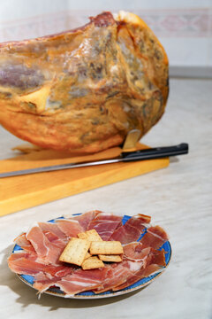 Serrano ham leg mounted on a jamonero with long carving knife, thin slices arranged on a plate beside crackers and wooden board, rustic spanish tapas presentation
