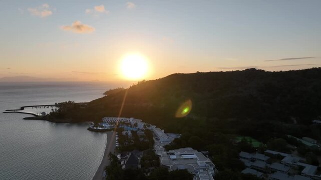Slow drone pullback with a gentle orbit at sunset, widening from Hayman Island resort and marina to the curved bay and open ocean with golden reflections, Whitsundays, Queensland, Australia.