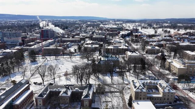 Penn State University in State College, Pennsylvania, with snow-covered campus buildings, frosted trees and surrounding hills under bright cold winter sky. Aerial wide shot panorama.