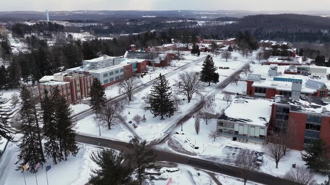 Aerial winter view of Saint Francis University in Loretto, Pennsylvania, showing snow-covered campus buildings, quiet roads and surrounding forested hills under cold gray sky. Wide shot.
