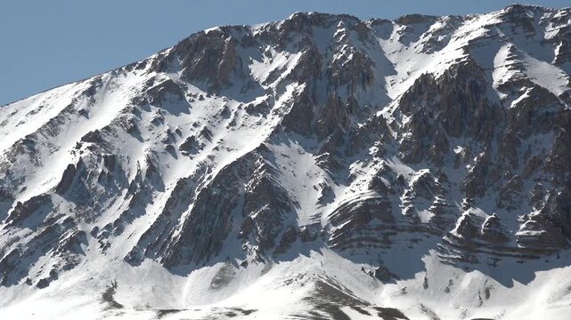 Rocky walls of a snowy dome mountain summit rise beneath a clear sunny winter sky. Dramatic alpine geology reveals rugged cliffs, frozen slopes, and pristine high elevation wilderness.
