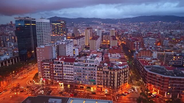 Drone pulling back over La Nova Esquerra de l&rsquo;Eixample at dusk, revealing illuminated streets, dense residential buildings and mountains beyond Barcelona.
