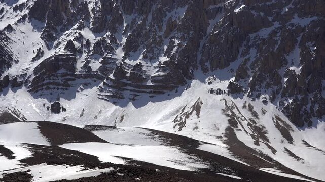 Rocky walls of a snowy dome mountain summit rise beneath a clear sunny winter sky. Dramatic alpine geology reveals rugged cliffs, frozen slopes, and pristine high elevation wilderness.