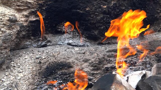 Natural methane gas fire burns between rocks on barren ground across a rugged rocky landscape. Eternal flames rise from stone gaps as subsurface fuel ignites within raw desolate terrain.
