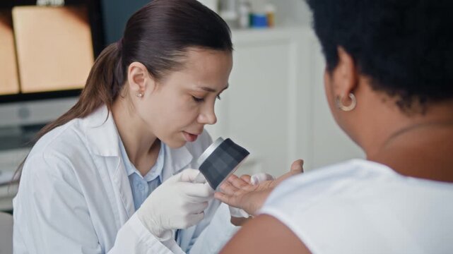 Professional dermatologist inspecting patient hand and fingers with dermatoscope during melanoma screening in modern clinic office, with closeup of skin mole on computer monitor in blurred background