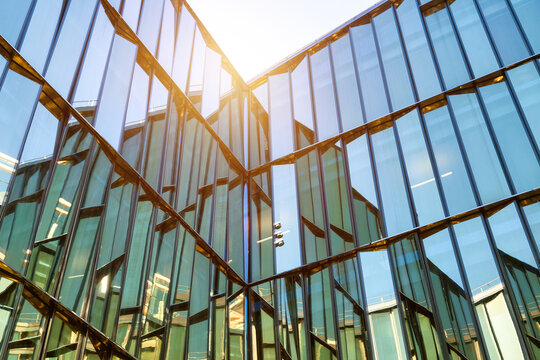 The sleek glass facade of a modern corporate building reflects a clear blue sky and bright sun during the day. Geometric patterns create an abstract background, offering ample copy-space.