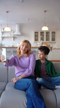 Charming blonde teenage babysitter and cute African American boy making video call online using mobile phone, chatting about day routine with mother while enjoying leisure at home.