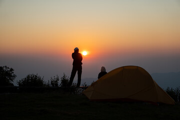 Silhouette of people camping with an orange tent on a high mountain peak enjoying a spectacular glowing sunrise. © artrachen
