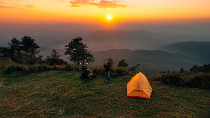 Aerial view of a male camper standing near an orange tent and motorcycle on a mountain ridge during a beautiful sunset. © artrachen