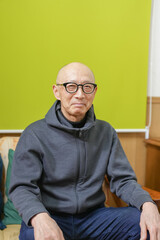 An old man wearing glasses and a gray hoodie sits on a couch. He looks sad and lonely