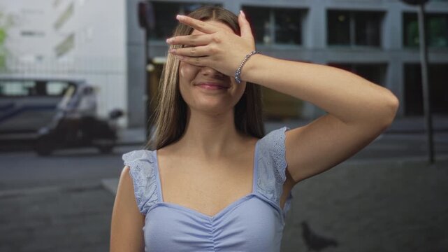Teenage girl wearing a light blue top raises hand to cover her eyes on a street with passing cars; shyness.