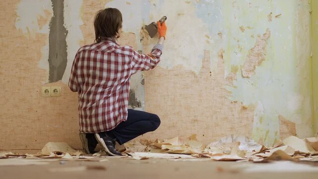 Young worker in protective gloves removing wallpaper from the wall during house repair and renovation. copy space