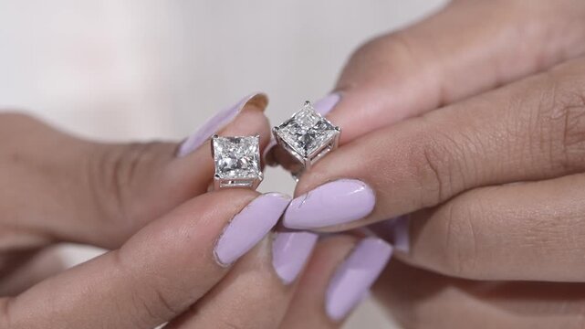 Macro close-up of a woman holding sparkling princess-cut diamond stud earrings between her fingers, showcasing elegant fine jewelry with a modern minimalist aesthetic on a soft neutral background.