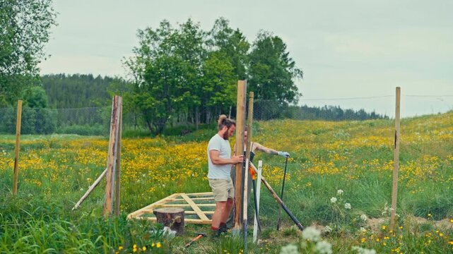 Static shot of a man extracting a wooden fence post as a woman assists him in a blooming meadow.