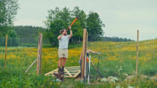 Static shot of a man and woman performing manual labor on a farm as a team to drive a wooden fence post into the ground using a splitting maul by a netted fence.
