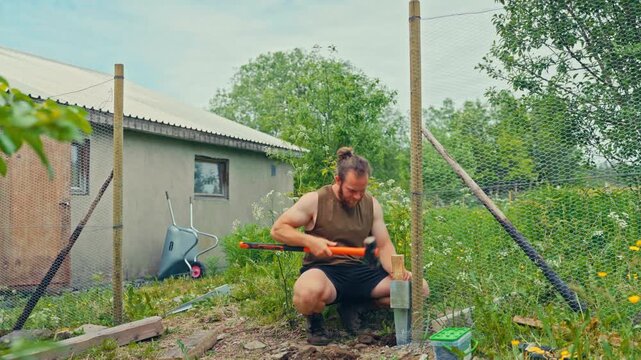 Static shot of a man using a sledgehammer to drive a metal post-spike in the ground as anchor for a wire-mesh netted fence.