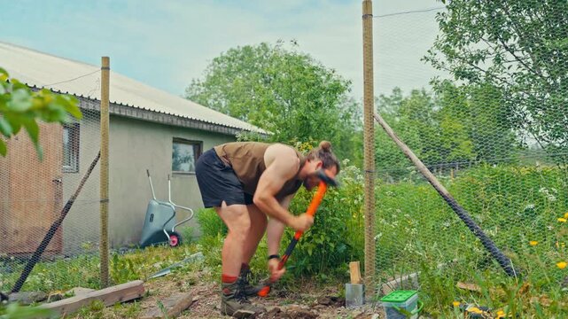Static shot of a man using a heavy splitting maul to hammer a wooden wedge into the base of a pole for a netted wire-mesh fence on a farm.