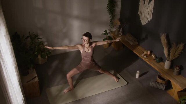 Wide overhead view of young experienced female yoga devotee performing warrior asana flow while exercising at home, in practice room adorned with green plants, deep shadows on walls