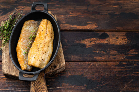 Crispy fried sea bass fish fillet with thyme in cast iron pan, mediterranean seafood dinner composition with rustic wooden background and copy space.