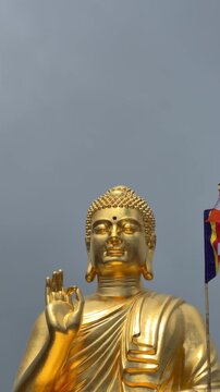 Vertical shot of a golden Buddha statue with Vitarka mudra raised hand, set against open sky and drifting clouds above the temple grounds. Buddhist gesture.