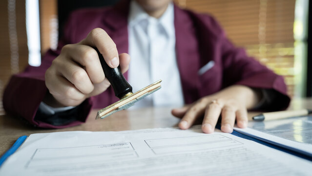 A person is signing a document with a stamp. The stamp is black and has a gold edge. The person is wearing a red suit and white shirt. The document is on a table