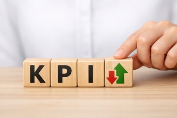 Naklejka premium KPI: Strategic Insights: A close-up view spotlights a hand artfully arranging wooden blocks spelling out KPI, paired with upward and downward arrows, representing key performance indicators.