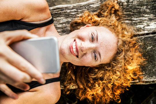 Portrait of redheaded young woman lying on bench using cell phone