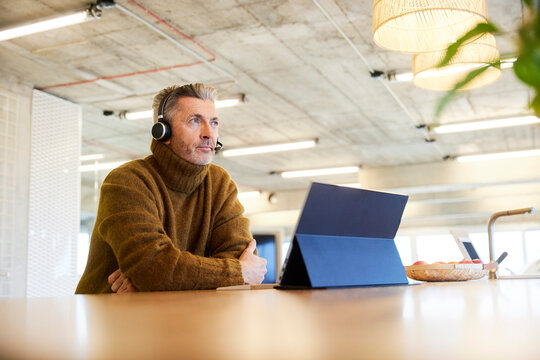 Thoughtful businessman with headset and digital tablet looking away while sitting at office