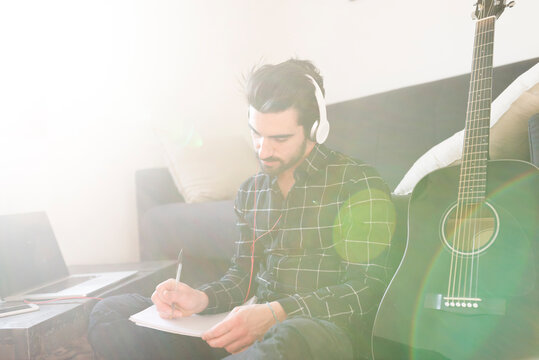 Young man at home with guitar wearing headphones and taking notes
