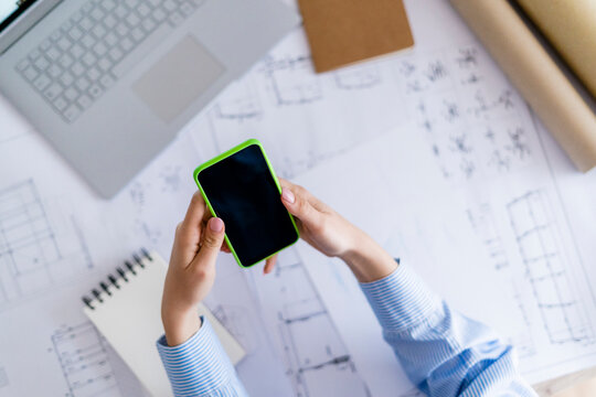 Close-up of woman in office using cell phone with blueprint on table