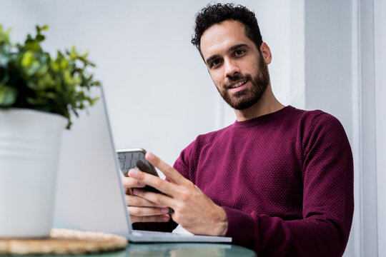 Portrait of young man stting at table at home using laptop and cell phone