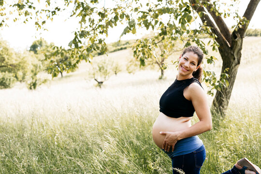 Young pregnant woman holding baby belly, standing on a meadow