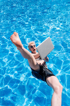 Shirtless young man holding laptop falling in swimming pool