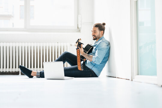Man sitting with laptop on floor playing guitar