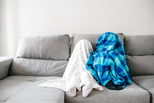 Little girl and her brother sitting side by side on the couch at home hiding under blankets
