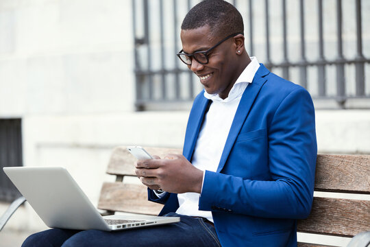 Young businessman wearing blue suit jacket sitting on bench and using smartphone