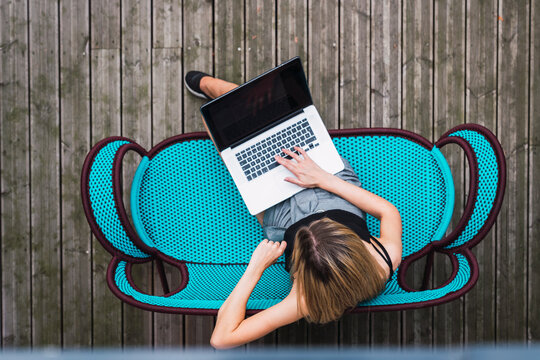 Young woman sitting on turquoise couch on terrace using laptop, top view