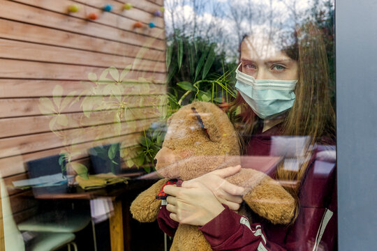 Portrait of girl with surgical mask and teddy bear behind window pane