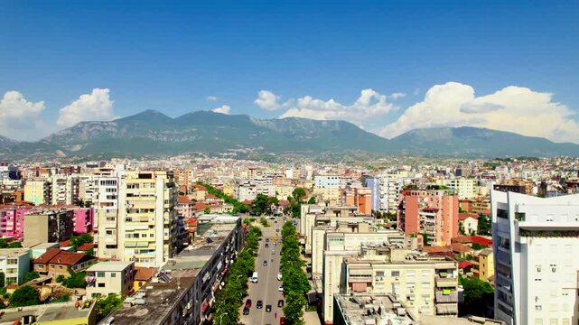High drone rise over Tirana, Albania, revealing roundabout and Rruga Luigj Gurakuqi leading toward Dajti Mountain. Panoramic cityscape with blue sky, clouds and summer atmosphere.