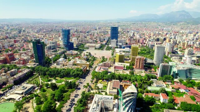 High drone flight over Tirana, Albania, approaching Skanderbeg Square and the National Historical Museum. Wide city skyline with mountains in background, summer daylight, green urban landscape.