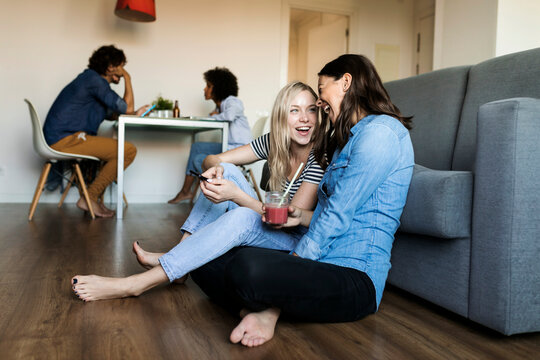 Two cheerful young women sitting on floor with cell phone and drink and friends in background