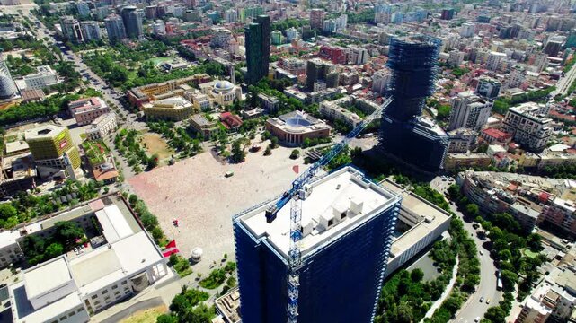 Drone orbit around a tall skyscraper under construction with rooftop crane in central Tirana, revealing Skanderbeg Square and surrounding modern high-rise buildings. Summer daylight, urban development