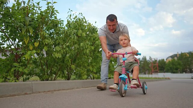 Happy father pushes little son on tricycle along street. Smiling man helps boy ride bike outdoors. Dad and child have fun together in summer. Family enjoys active leisure time on sunny day.