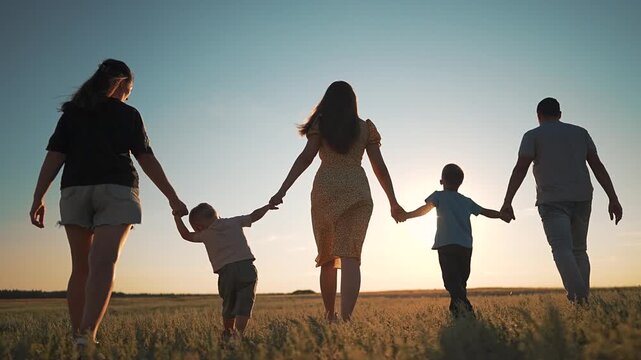 Big happy family is walking in green field at sunset. Parents and children hold hands in a row. Group of people enjoys summer evening in nature. Large family walks away together in park happily.