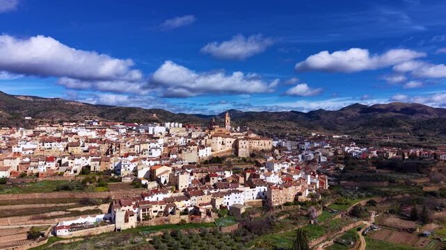 Aerial panorama of Chelva, showcasing historic stone architecture, terraced hillsides, and mountains beneath an epic mix of sunlight and fast-moving clouds